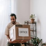 Man carrying a medium cardboard box labeled Office through a bright room filled with packed moving boxes