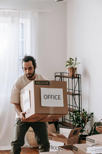 Man carrying a medium cardboard box labeled Office through a bright room filled with packed moving boxes